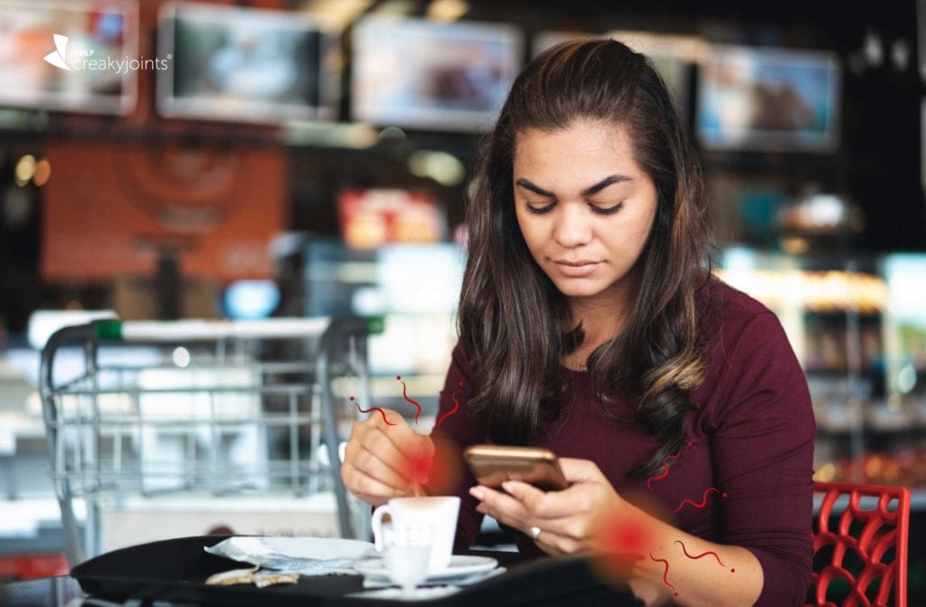 Photo shows a Hispanic woman in a coffee shop using her cellphone. There are red spots on her wrists indicating joint pain