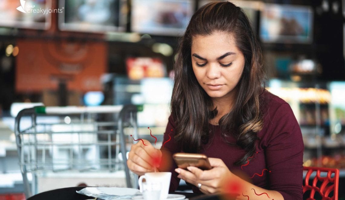 Photo shows a Hispanic woman in a coffee shop using her cellphone. There are red spots on her wrists indicating joint pain