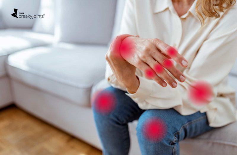 Photograph shows a woman sitting on a couch. She has red spots indicating joint pain