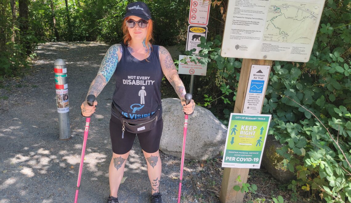 A photo of a woman, Eileen Davidson, standing in front of the entrance to a hike. She is holding hiking poles.