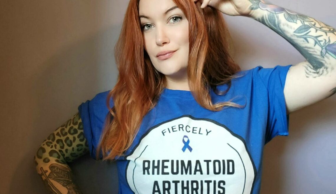 A photo of a woman, Eileen Davidson, modeling the "Fiercely Fighting Rheumatoid Arthritis" t-shirts, which will be available for purchase following the Tumbler and Tipsy by Michael Kuluva 2022 Collection event, airing on Tuesday, September 14, 20201 at 8 p.m. ET via YouTube. Eileen is wearing a blue shirt with a white circle outlined in black. Within the white circle are the words "Fiercely." Beneath that is a blue ribbon to symbolize arthritis awareness. Below the ribbon are the words "Rheumatoid Arthritis." Below those words are an image of a hand in a fist with lightning bolts. Below the hand is the worth "Fighting.