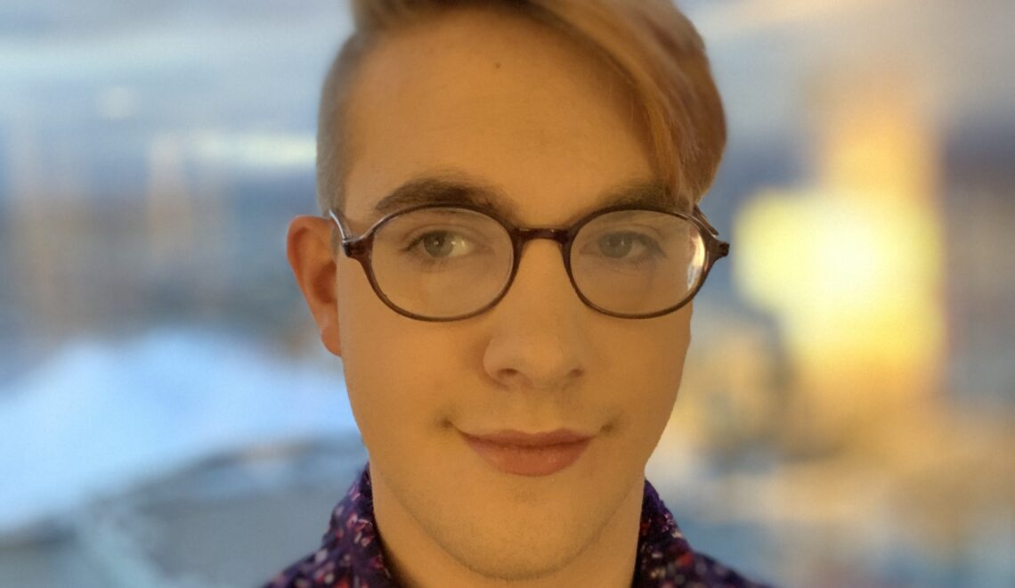 Derek is wearing a purple speckled button-down, a light red lip, base makeup, and their glasses. Their hair is fluffed to the right. The image has a blurred gloomy San Diego skyline in the background.